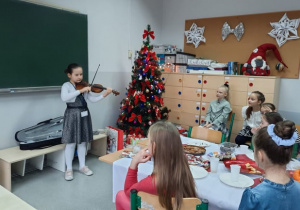 Na zdjęciu widać uczennicę, która gra na skrzypcach. Podziwiają ją inni uczniowie, siedzący przy stole. W tle widać choinkę.