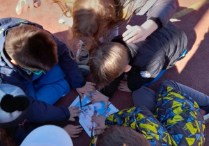 Na zdjęciu widac uczniów znajdujących się na boisku szkolnym, pochylonych nad kartką, wykonujących zadanie.