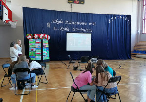 Na fotografii widać uczniów, którzy biorą udział w quizie na temat świąt majowych. W tle znajduje się ekran oraz dekoracje.