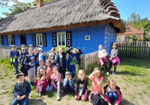 Na zdjęciu widać uczniów, w tle znajduje się niebieski domek wiejski w skansenie.